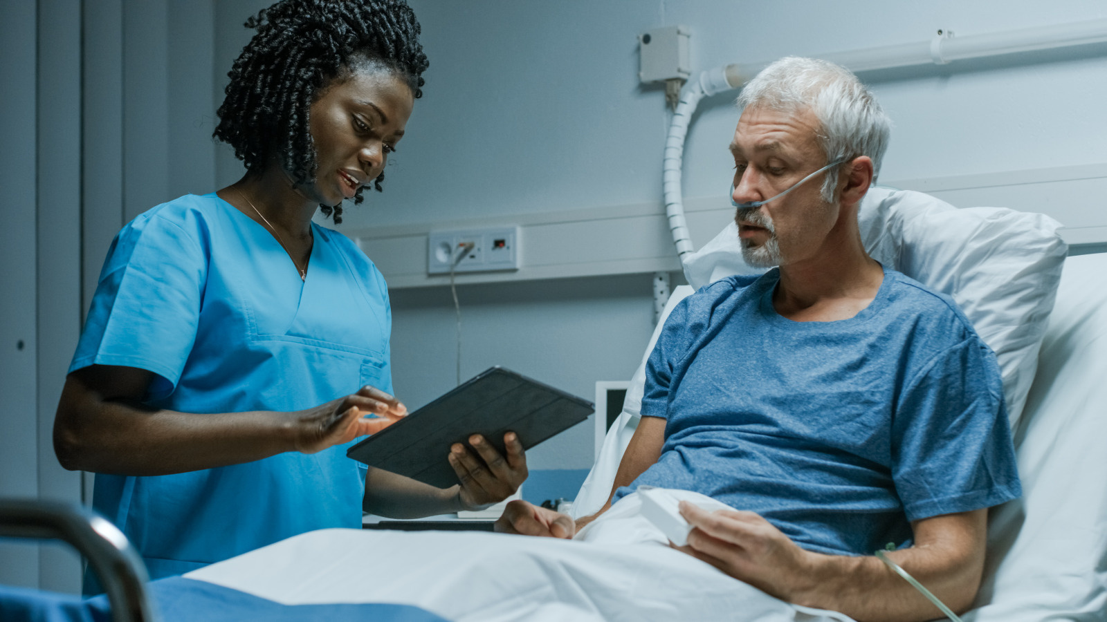 Jeune infirmiere tenant une tablette et un malade alite a l'hopital - Infirmier-e en pratique avancee